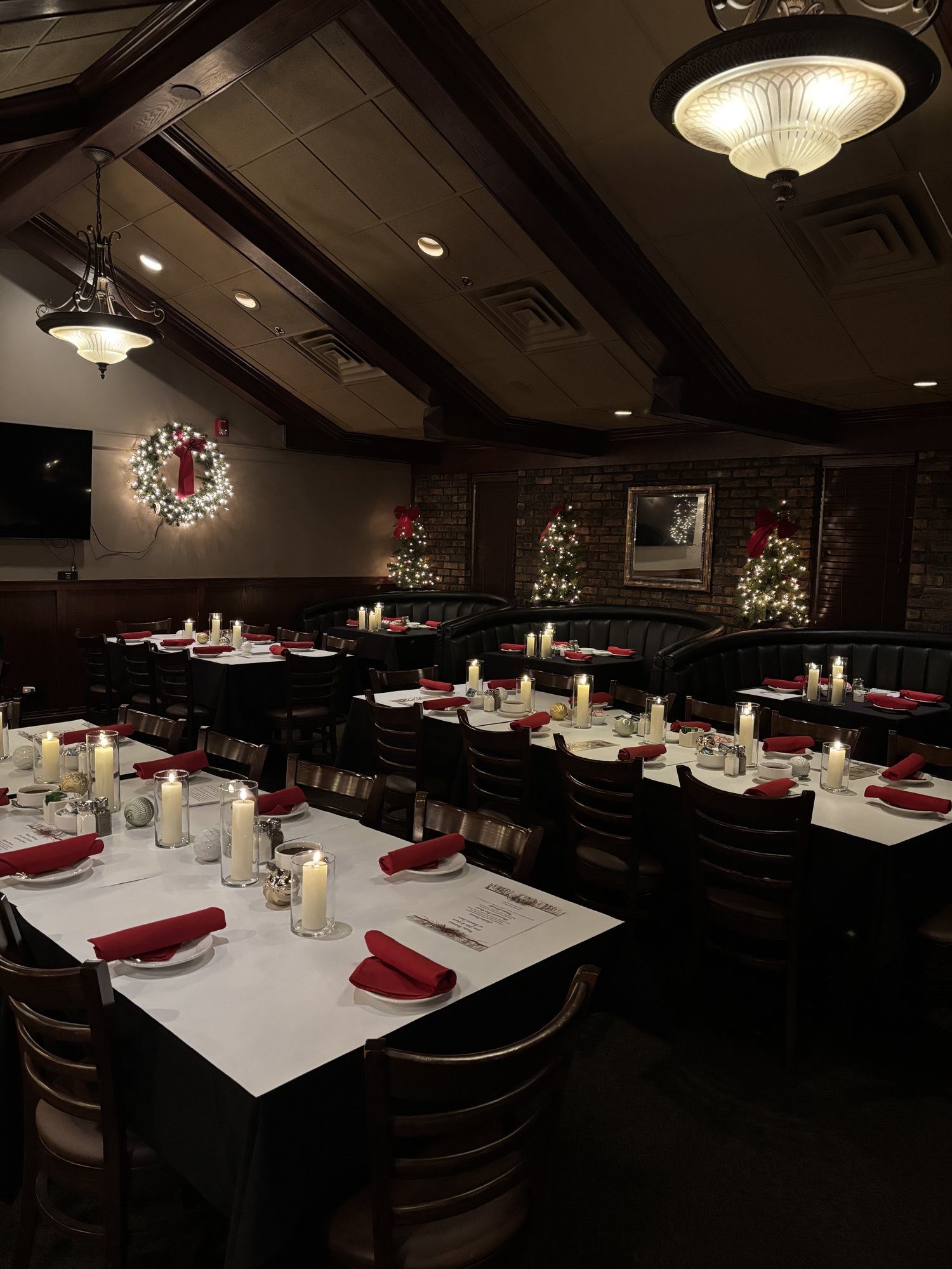 A private dining room dressed for a holiday party with candlelit tables, red napkins, a lit wreath and three decorated Christmas trees.