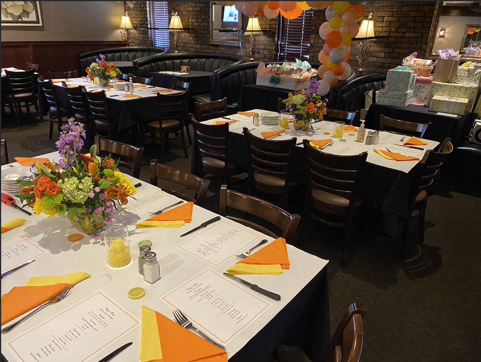 A private room set for a baby shower with white linens, orange and yellow napkins, floral centerpieces, a balloon arch and a gift table.
