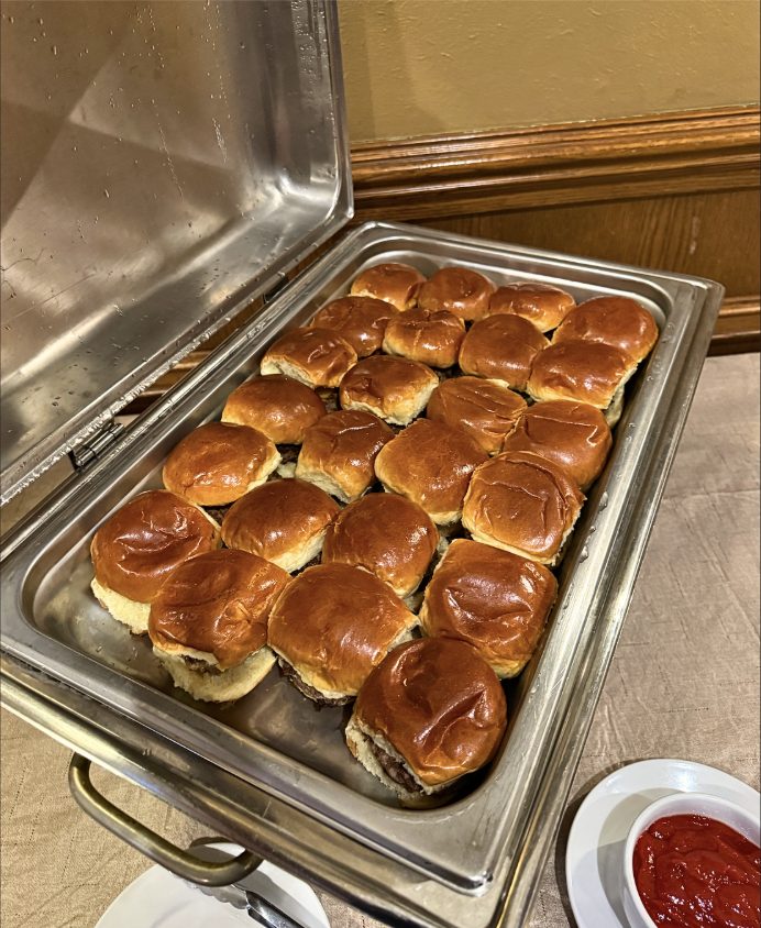 A chafing dish of mini burger sliders on brioche buns with a bowl of ketchup.