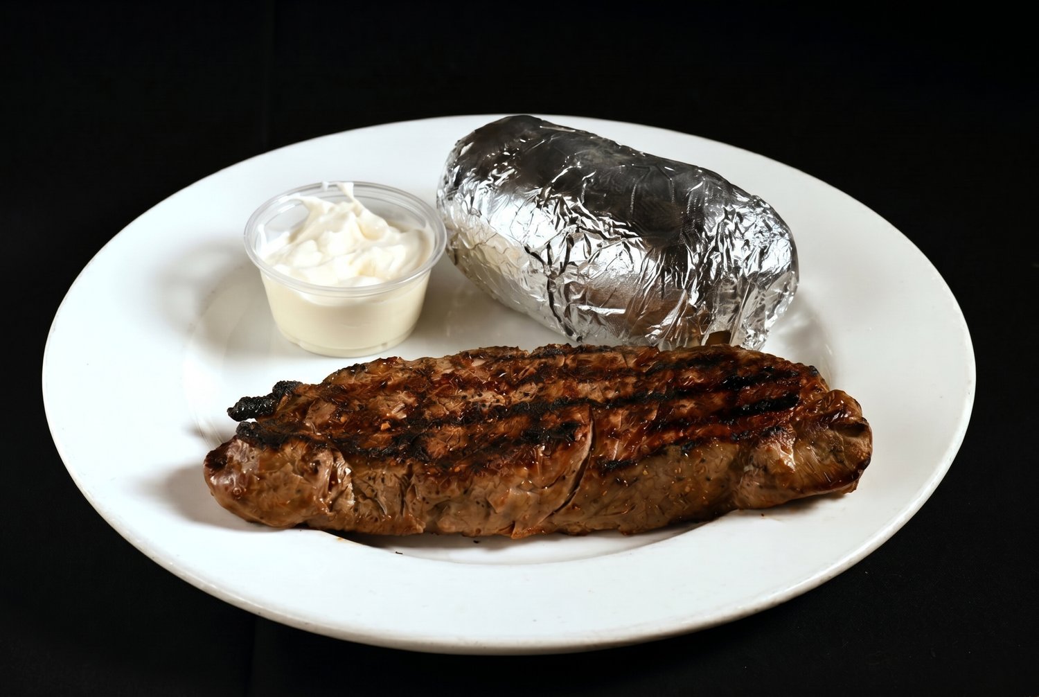 Char-grilled New York strip steak with grill marks, served with a foil-wrapped baked potato and sour cream.