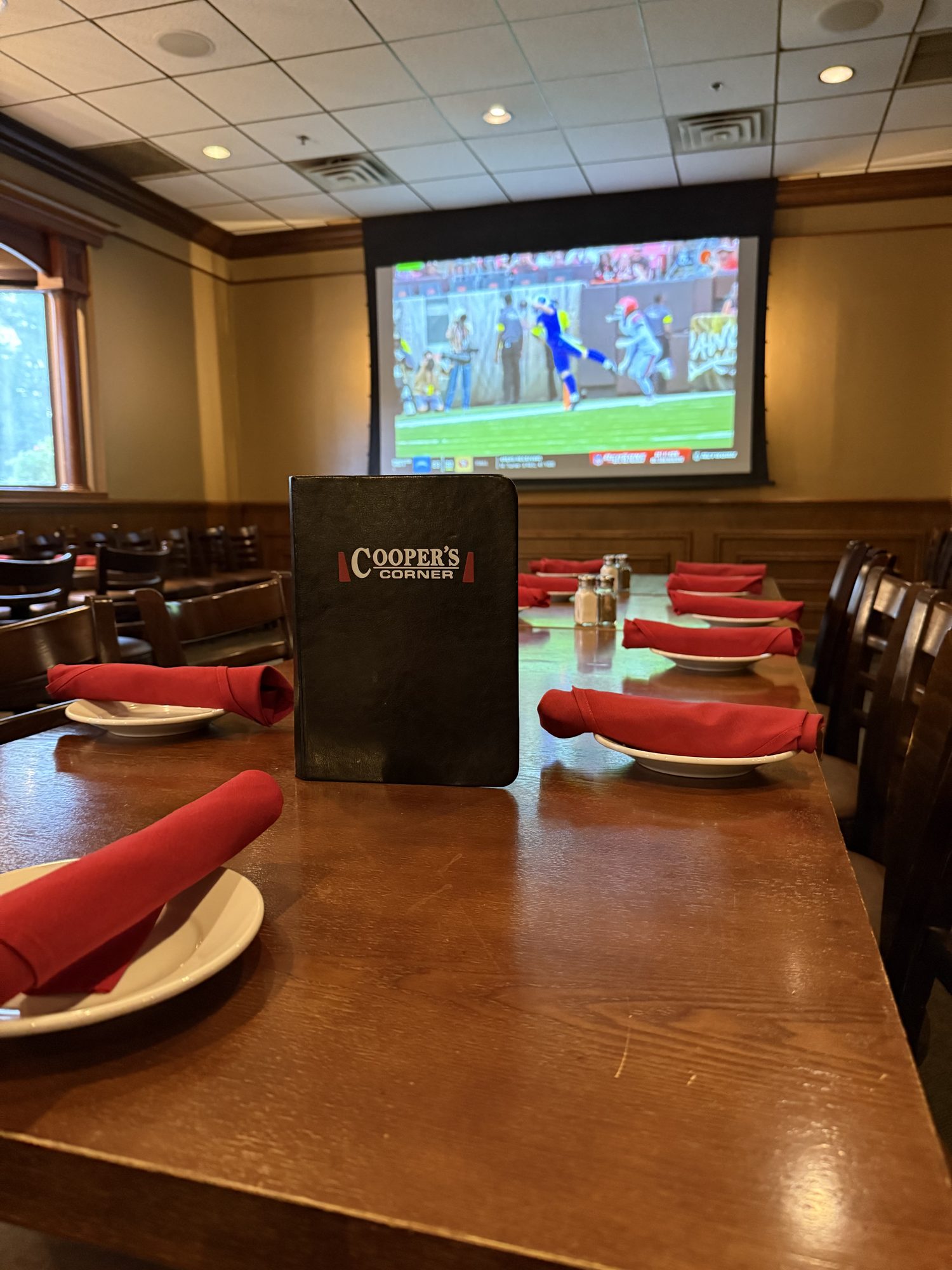 A long banquet table set with red napkins facing a 120 inch projection screen showing a football game.
